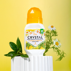 Crystal deodorant bottle with orange cap on yellow background, surrounded by green leaves and white daisies.