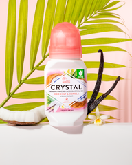 Crystal mineral-enriched deodorant roll-on bottle with pink faceted cap, coconut and vanilla scent, framed by tropical palm leaves.