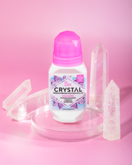 Crystal mineral enriched deodorant roll on bottle with a pink gem cap surrounded by clear crystals against a pink backdrop