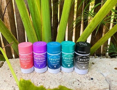 Assorted Crystal deodorant bottles with vibrant caps arranged in a row on a stone surface with leafy plants in the background.