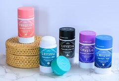 Crystal mineral deodorants in pink teal black purple and blue bottles arranged on a marble surface beside a woven basket.