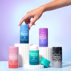 Cluster of Crystal deodorant bottles in different colors arranged on a white surface against a pastel gradient backdrop.